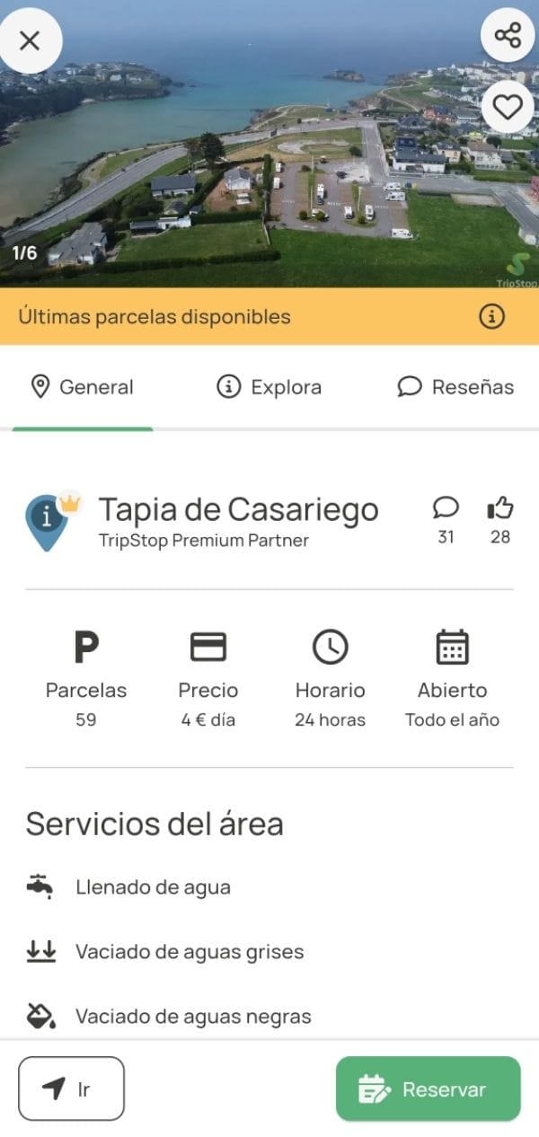 Vista aérea del área de autocaravanas de Tapia de Casariego, con parcelas delimitadas, casas cercanas, acceso por carretera y el mar al fondo.