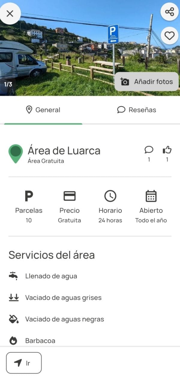 Zona de aparcamiento para autocaravanas en Luarca, con señal de estacionamiento, césped, y vistas al pueblo sobre una colina.