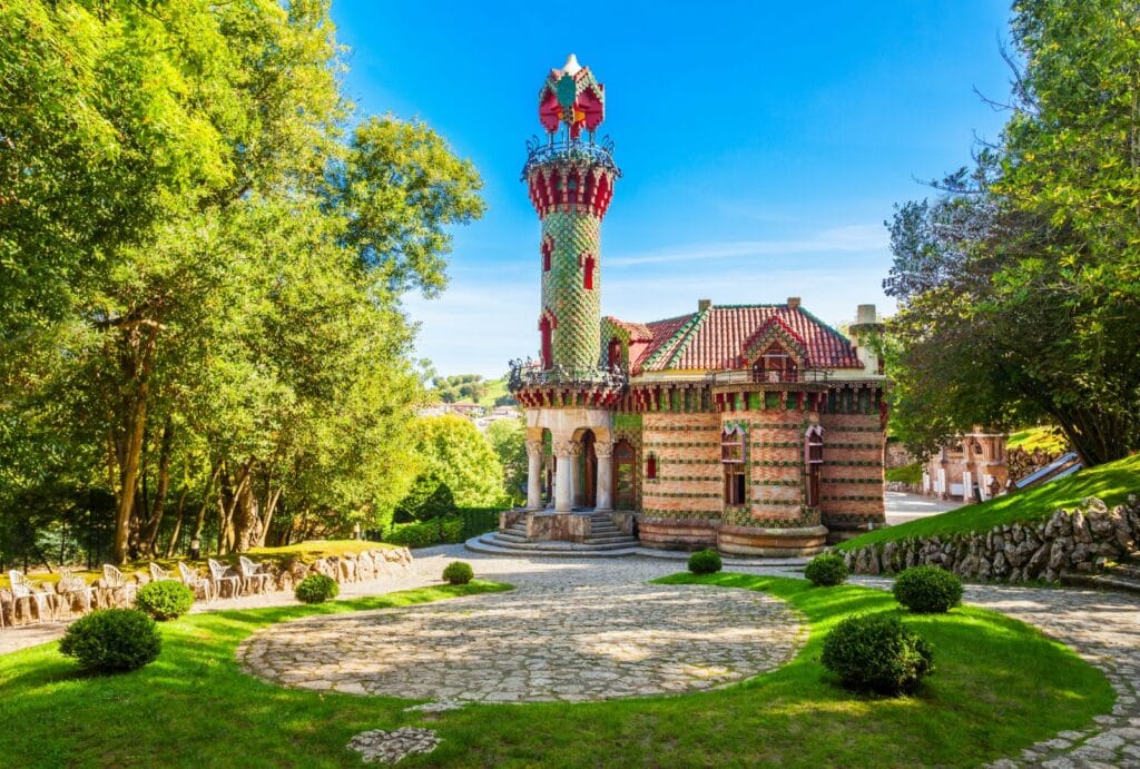 Edificio modernista conocido como El Capricho de Gaudí, con una torre cilíndrica decorada con cerámicas verdes y rojas, rodeado de jardines verdes y árboles bajo un cielo azul despejado en Comillas, Cantabria.