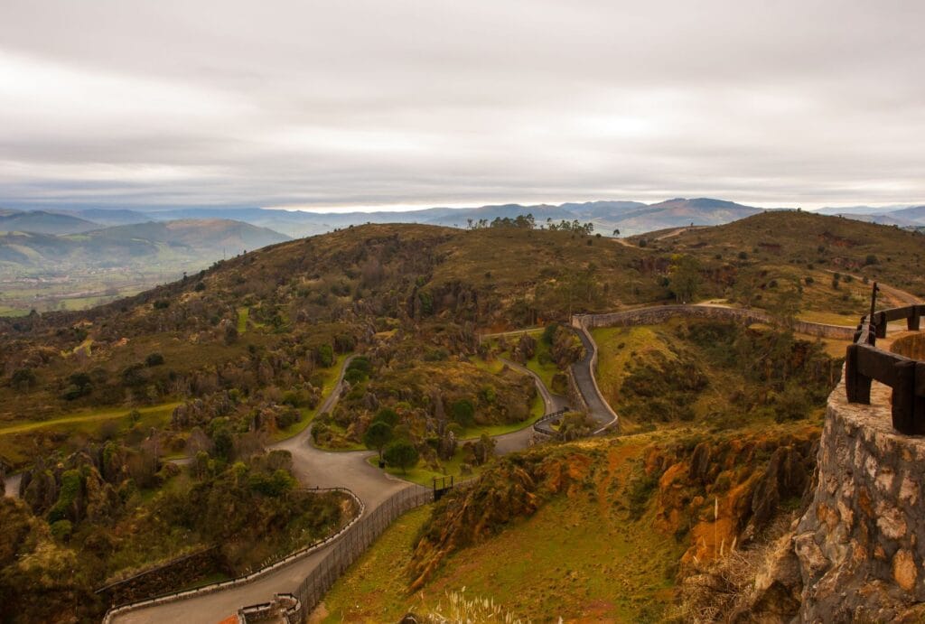 Vista panorámica de un paisaje montañoso con caminos serpenteantes, vegetación y cielo nublado, desde un mirador de piedra en Cantabria.