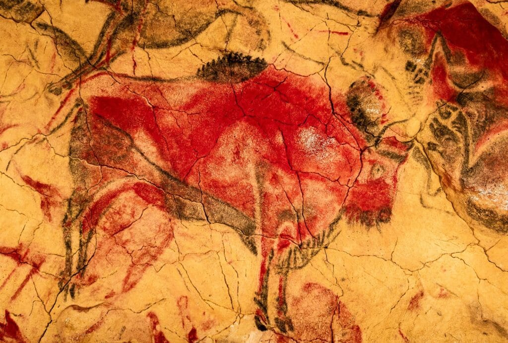 Pintura rupestre de un bisonte en tonos rojos y negros sobre fondo ocre, ubicada en una cueva prehistórica con grietas visibles en la pared rocosa.