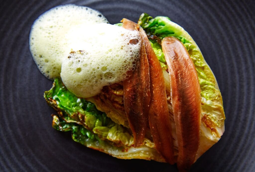 Plato gourmet con hojas de lechuga a la plancha, anchoas del Cantábrico y espuma blanca servidos sobre un plato oscuro.