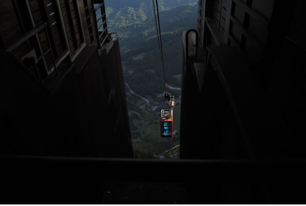 Teleférico rojo descendiendo desde una estación de alta montaña entre dos estructuras oscuras, con vista al valle verde de fondo.