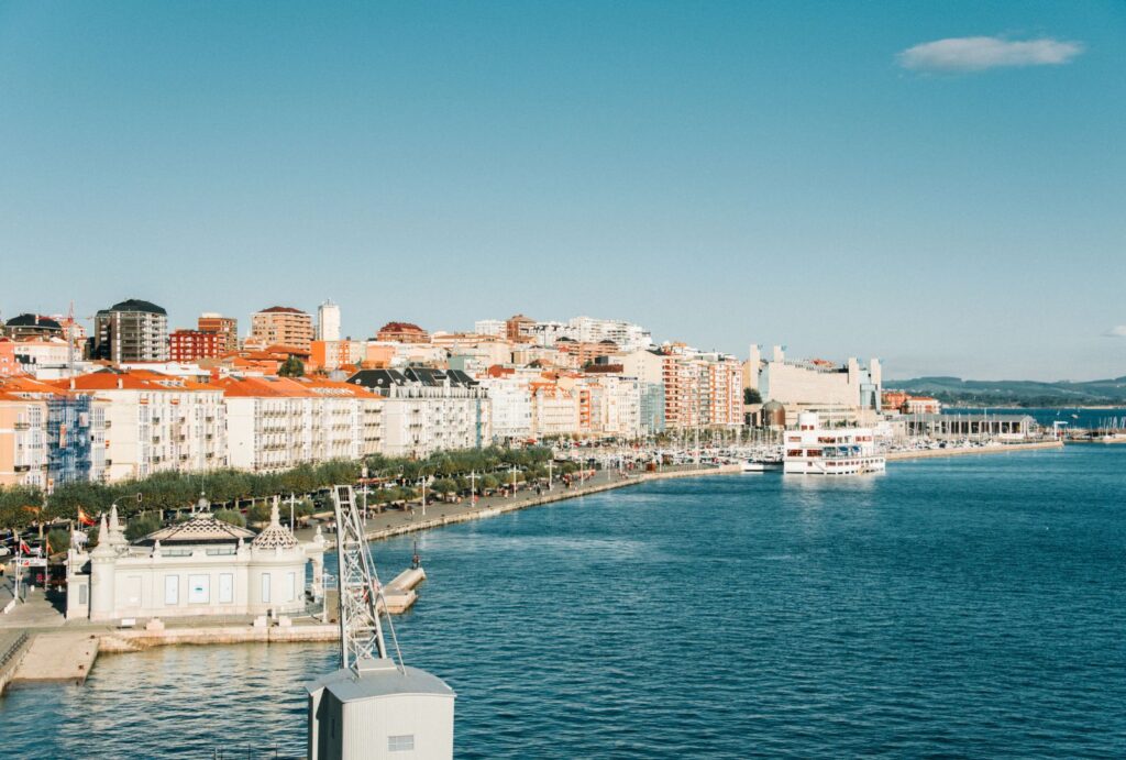 Vista panorámica del paseo marítimo y el puerto de Santander con edificios coloridos, un barco turístico y cielo despejado.