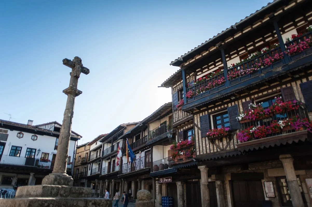 Imagen de una plaza de pueblo en La Alberca, España, con casas de arquitectura tradicional de madera y piedra adornadas con balcones llenos de flores de colores vivos.