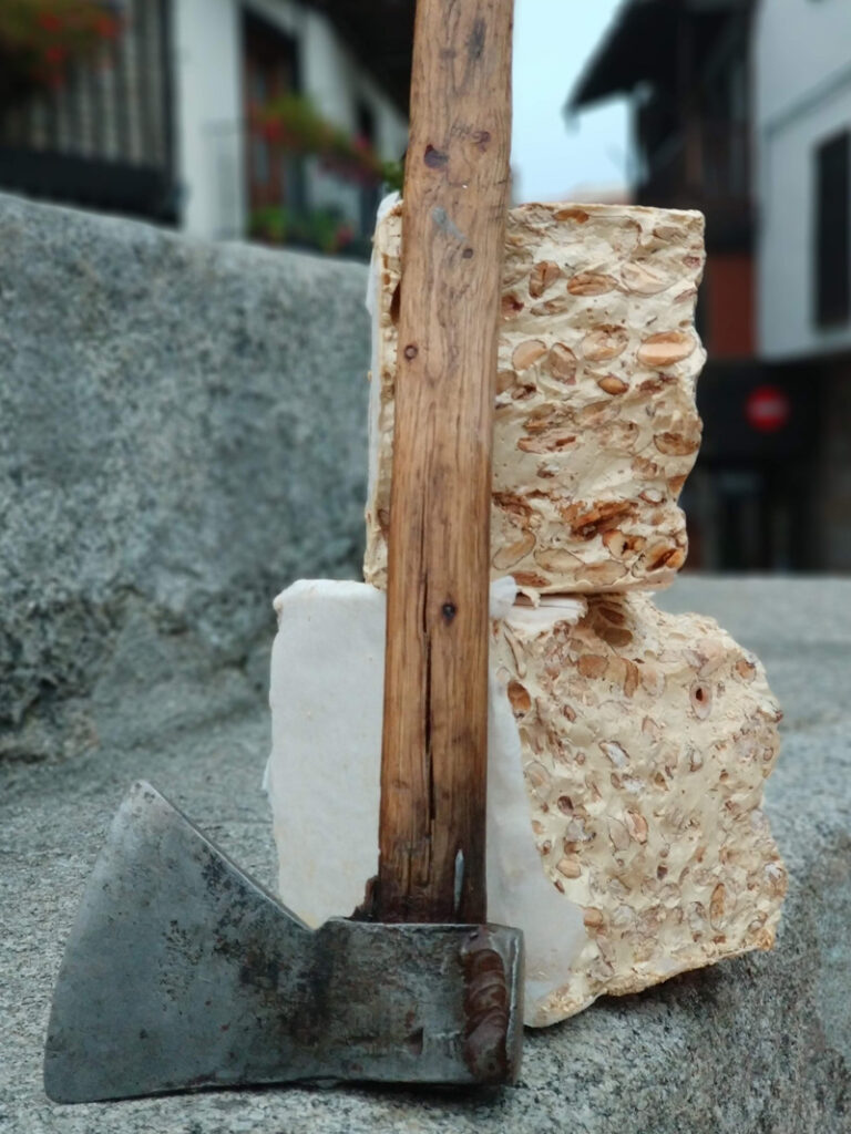 Primer plano de un trozo grande de turrón duro con almendras apoyado sobre un escalón de piedra, junto a una vieja hacha de madera y metal.