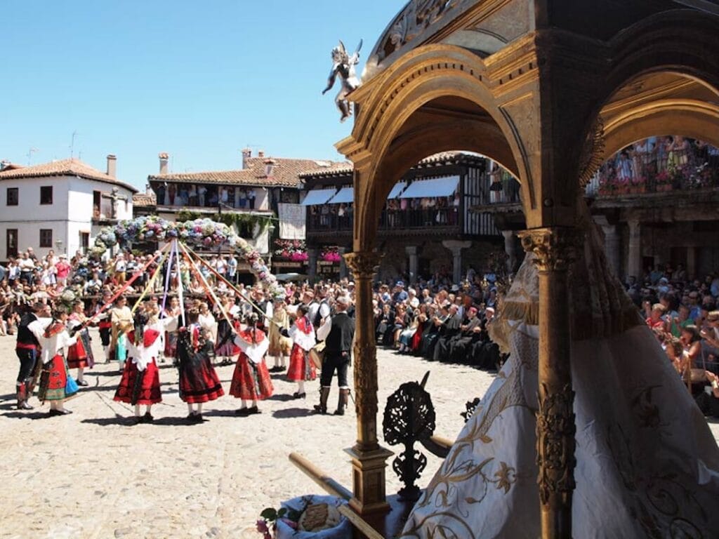 Celebración tradicional en La Alberca con mujeres y hombres vestidos con trajes típicos charros bailando alrededor de un arco floral con cintas de colores en la plaza del pueblo, mientras la imagen de la Virgen de la Asunción observa desde un paso dorado en primer plano.