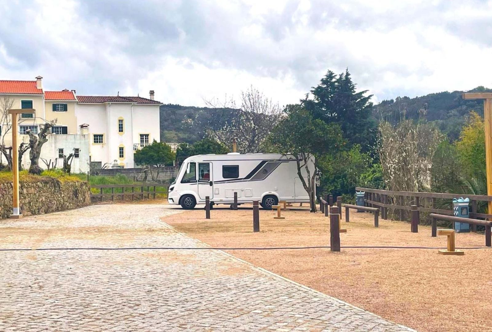 Autocaravana blanca estacionada en un área de descanso pavimentada con casas y colinas al fondo.