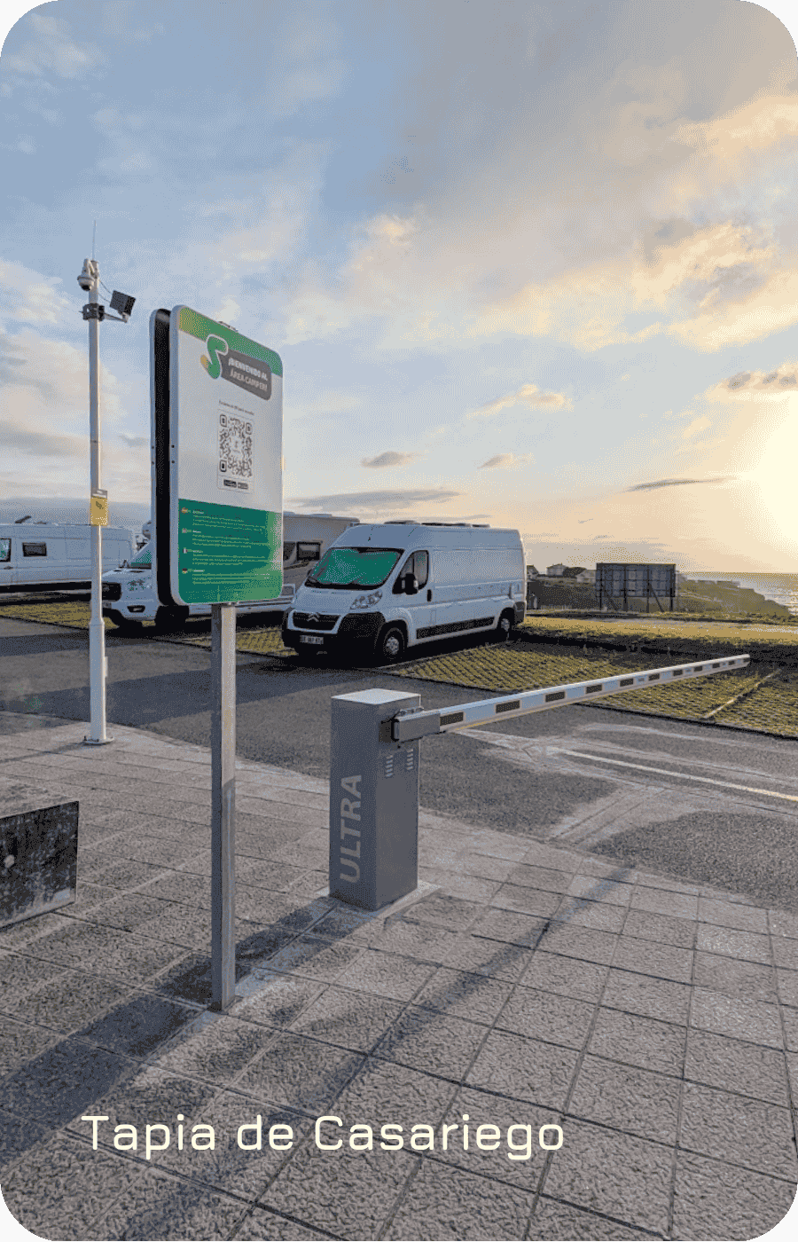 Entrada del área de autocaravanas de Tapia de Casariego. Se puede ver una barrera, un cartel con un QR, cámaras de seguridad y de lectura de matrículas. También se ven algunas autocaravanas al fondo y una bonita puesta de sol.