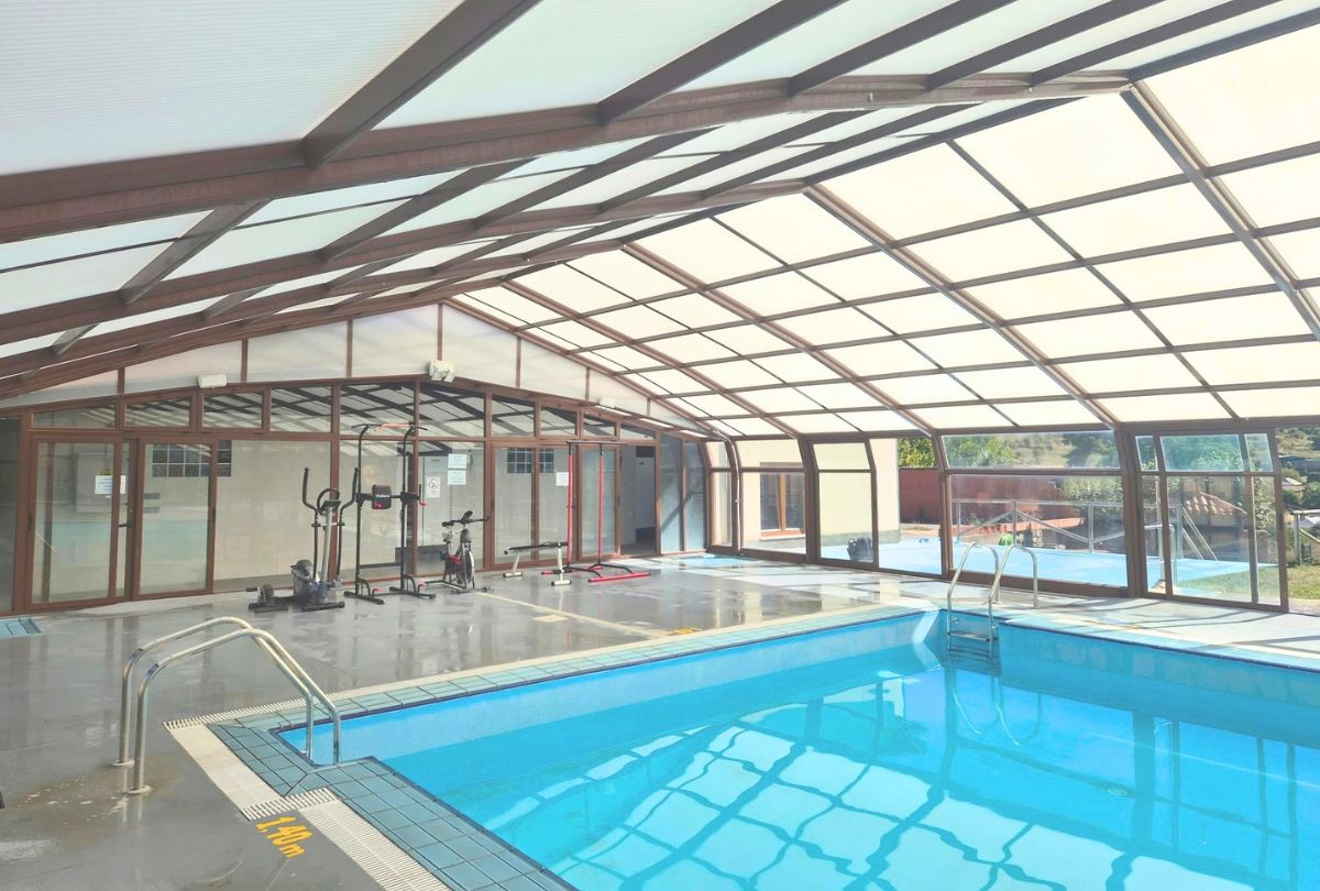 Piscina cubierta con techo translúcido de estructura marrón y grandes cristaleras abiertas al paisaje. Al fondo hay una zona con bicicleta estática, elíptica y otros aparatos de gimnasio, y en el borde se lee la marca de profundidad de 1,40 m.
