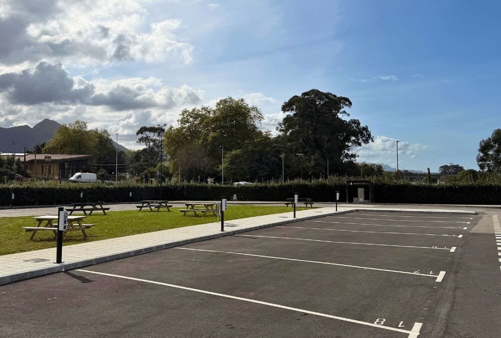 Aparcamiento en Llanes delimitado para autocaravanas sobre asfalto, con bornes de electricidad, zona verde con mesas de picnic de madera y árboles al fondo.