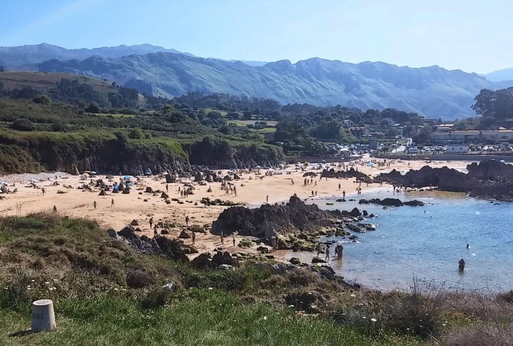 Vista panorámica de la playa de Toró en Llanes con bañistas entre sus características rocas en la arena, con colinas verdes y montañas de fondo en un día soleado