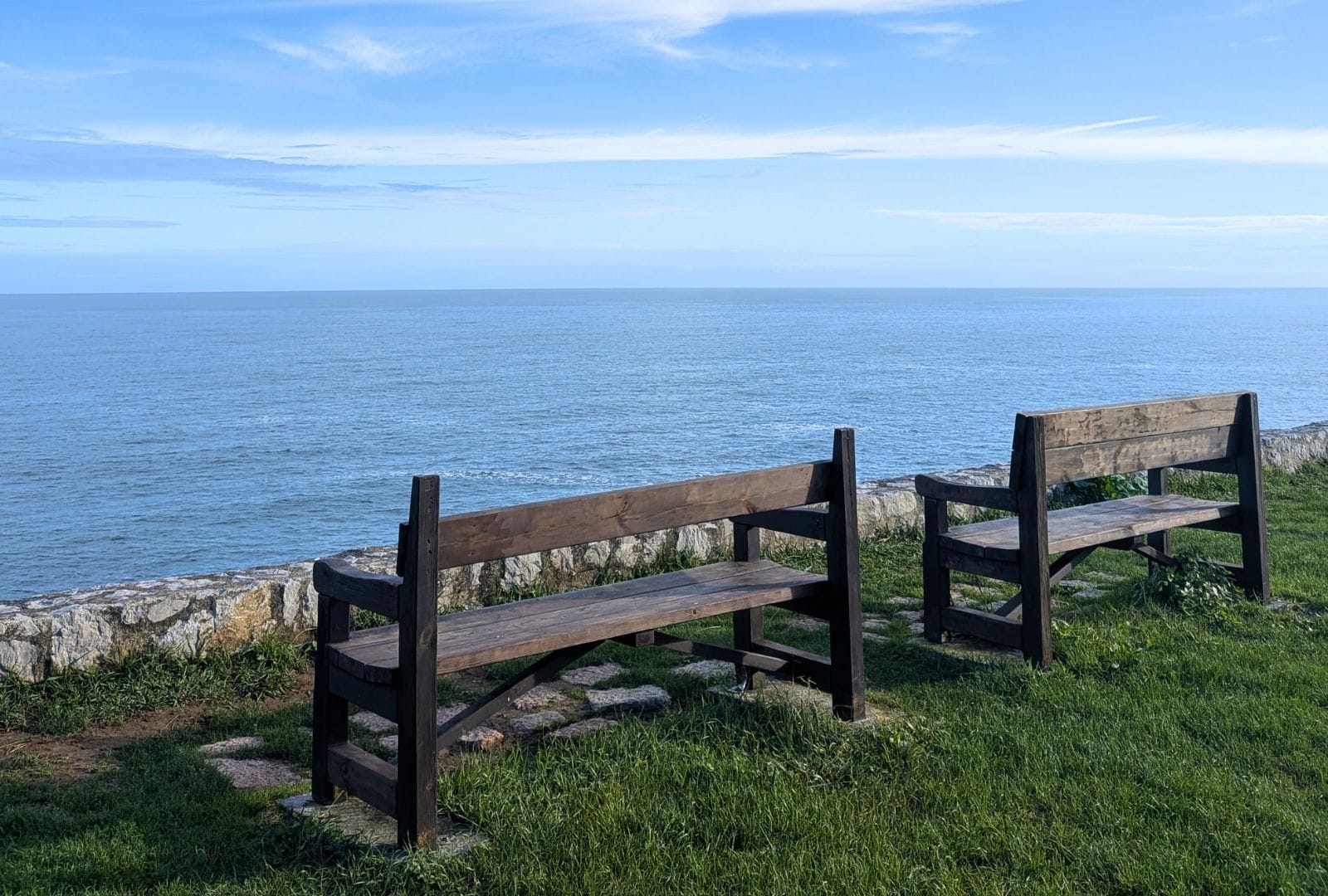 Dos bancos de madera sobre la hierba de un mirador junto al mar Cantábrico, con un murete de piedra y el horizonte despejado en la costa norte de España.